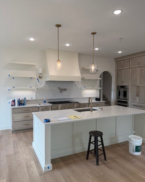 A nearly finished kitchen with a large white island, custom range hood, pendant lights, and light wood cabinets.