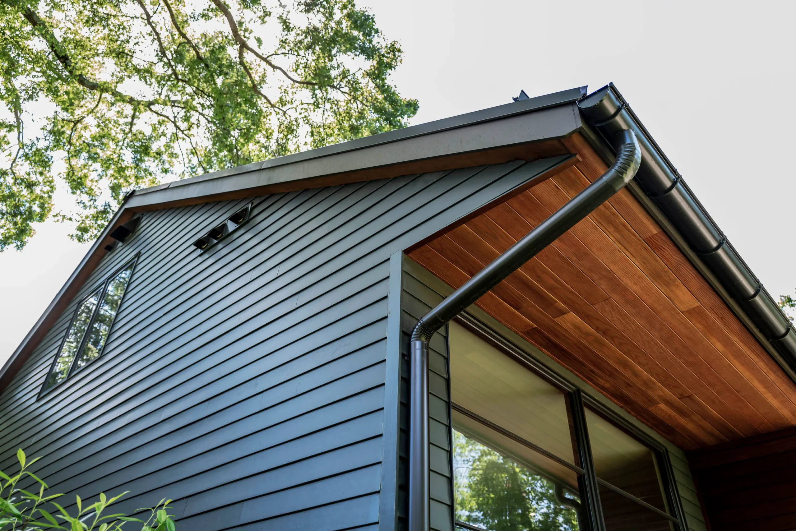 Close-up of a home roofline with gray lap siding, a dark metal gutter, and a warm wood soffit overhang.