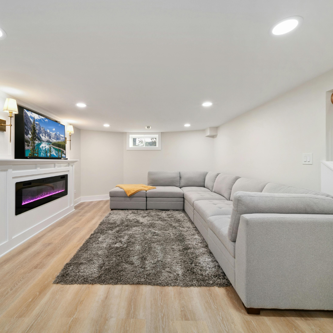 A finished basement with a gray sectional sofa, electric fireplace, wall-mounted TV, and light hardwood floors.