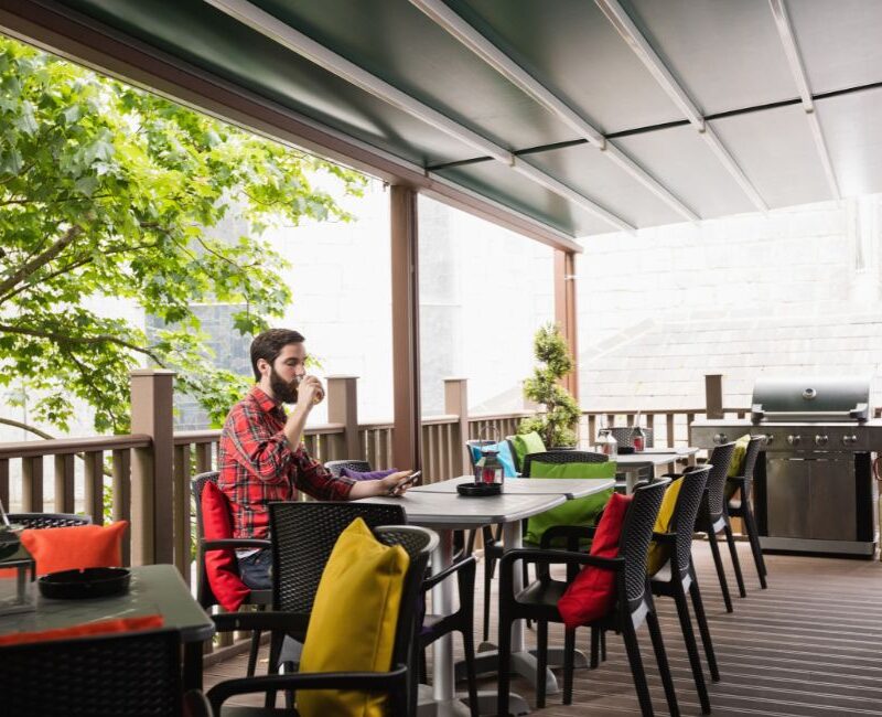 A covered deck with outdoor dining furniture, a gas grill, and a retractable awning surrounded by trees.