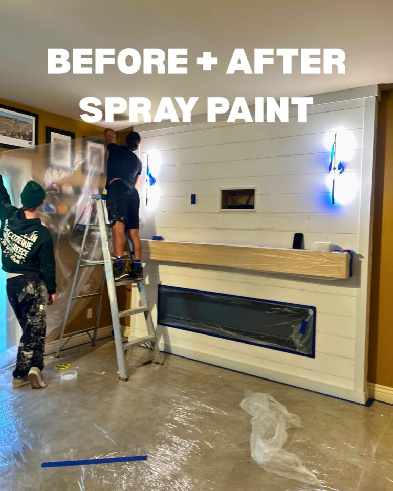 Two painters spray painting a shiplap accent wall with fireplace and wood mantel during a basement remodel.