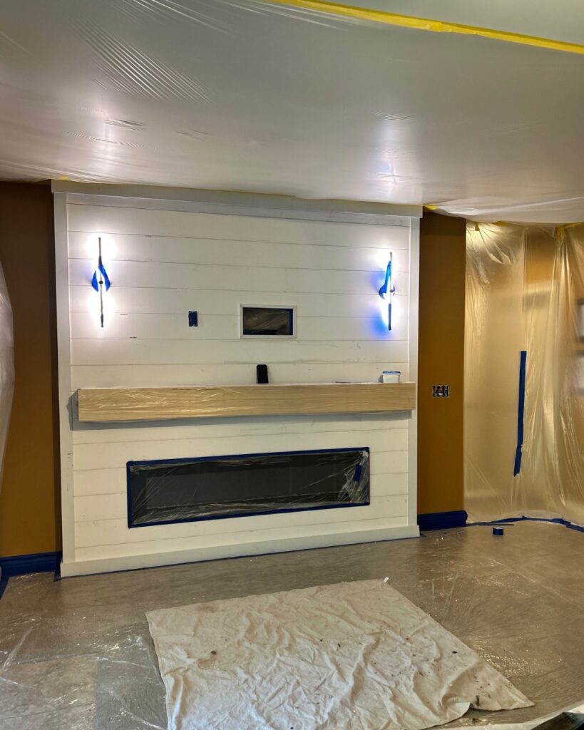 A shiplap accent wall with electric fireplace opening and wood mantel mid-construction before final painting.